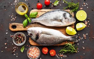 Horizontal image of two whole fish on a cutting board surrounded by tomatoes, herbs, citrus, and assorted seasonings.