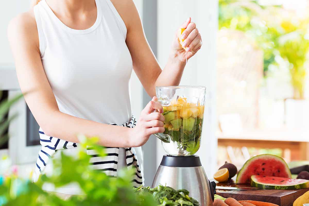 Horizontal image of a woman making a fresh shake in the kitchen on a counter next to sliced fresh fruit and vegetables.