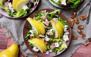 Horizontal top-down image of three dishes on a tan towel with a mix of baby greens, nuts, diced shallots, and sliced fruit.