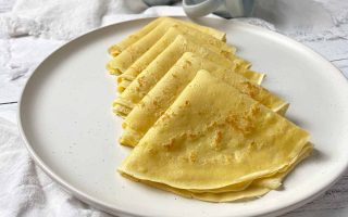Horizontal image of a row of folded thin pancakes on a white plate surrounded by white towels, with mugs in the background.