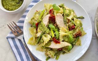 Horizontal image of a white dinner plate with farfalle mixed with chicken slices and assorted vegetables in a green sauce next to a fork and napkin.