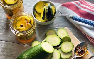 Horizontal image of slices of green vegetables on a wooden cutting board next to jars of pickles and a towel.