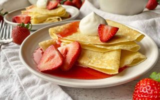 Horizontal image of two white plates, each with with a stack of folded thin pancakes served with whipped cream, sliced fruit, and a red syrup.