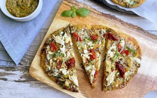 Horizontal image of three slices of an oblong pizza on a wooden cutting board next to pesto and a blue towel.