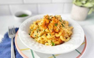 Horizontal image of a white bowl filled with elbow pasta in a cream sauce topped with toasted breadcrumbs and herbs next to a fork, blue towel, and a bowl of more chopped herbs.