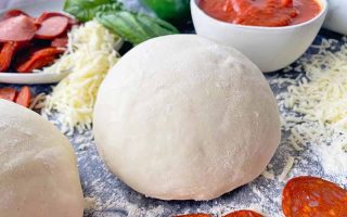 Horizontal image of a mound of lightly floured dough next to shredded cheese, basil, pepperoni slices, and a bowl of marinara sauce.
