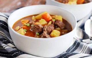 Horizontal image of white bowls filled with a soup with chunks of meat and vegetables on a striped napkin.