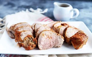 Horizontal image of slices of cooked seasoned meat on a white plate next to a cup of gravy and a towel.