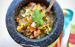 Horizontal image of a stone bowl filled with chopped vegetables topped with a fresh herb and a spoon inserted into it.