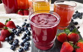 Horizontal image of three glasses filled with assorted drinks on a dark slate next to fresh fruit.