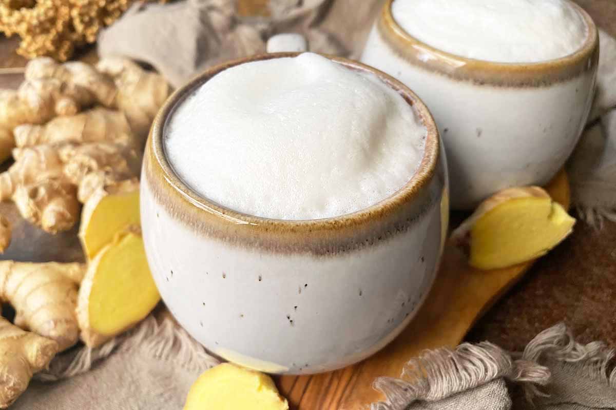 Horizontal image of two mugs on a wooden board filled with a foamy beverage next to sliced ginger.