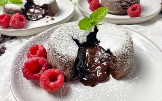 Horizontal image of three plates with chocolate cakes cut in half to reveal a gooey center, with raspberries and mint leaves as garnishes.
