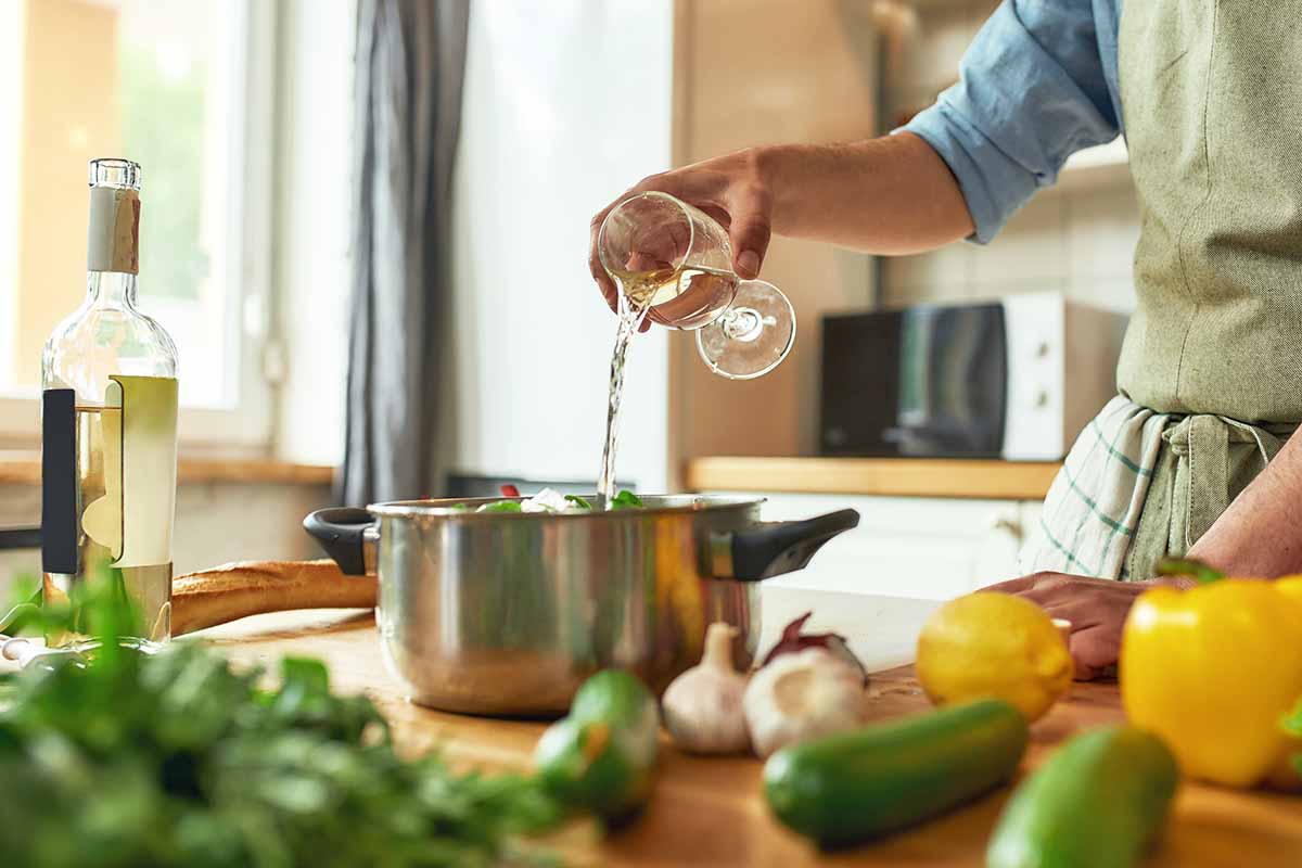 Horizontal image of a chef pouring wine into a pot with chopped vegetables while preparing a meal.