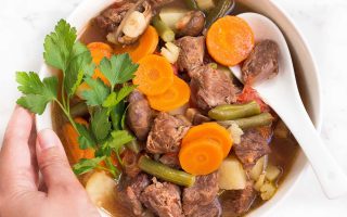 Horizontal image of a hand holding the side of a bowl full of a hearty meat and vegetable soup with a spoon.
