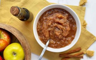 Horizontal top-down image of a white bowl filled with a cinnamon fruit spread on a yellow towel with a spoon.