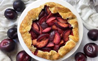 Horizontal top-down image of a large crostata with red fruit wedges next to a bowl of vanilla ice cream and metal forks on a white towel.