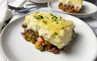 Horizontal image of a large square of a baked casserole topped with parsley on a white plate next to a white towel and metal forks.