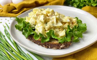 Horizontal image of an open-faced sandwich topped with lettuce and a creamy mix of hard-boiled yolks and whites on a white plate next to chives and a yellow towel.