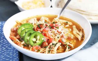 Horizontal image of a white bowl with a stew that has shredded meat, garnished with slices of peppers and a small mound of salsa.