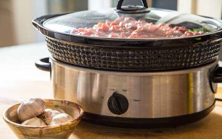 Horizontal image of an appliance filled with a stew on a wooden table next to a bowl of garlic.