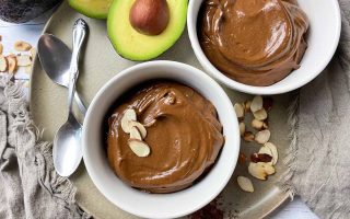 Horizontal top-down image of two white bowls filled with a creamy cocoa dessert, one topped with sliced almonds, on a plate next to a halved avocado and spoons.