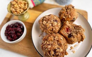 Horizontal image of a white plate with four oat-topped muffins next to bowls of dried fruit and nuts.